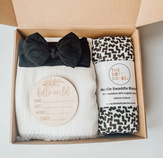 A gift box for a new baby with black and white tones. Featuring a black headband, white tassel blanket, hello world wooden disc and black zig zag swaddle.